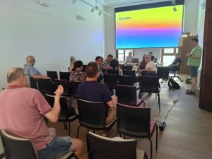 Fotografia dels membres de Sofcatalà a la trobada de pencaires d'estiu, d'esquena, asseguts davant d'una pantalla de projector, mentre un altre membre plantat fa una explicació.